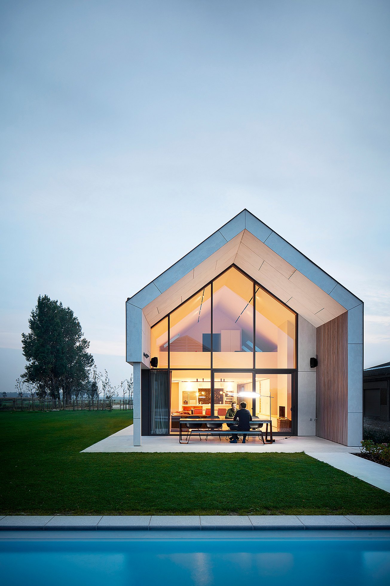 Une table Hopper et des chaises longues noires sont installées sur la terrasse d'une maison moderne à pignon, au crépuscule. On aperçoit la piscine à travers la façade vitrée illuminée.