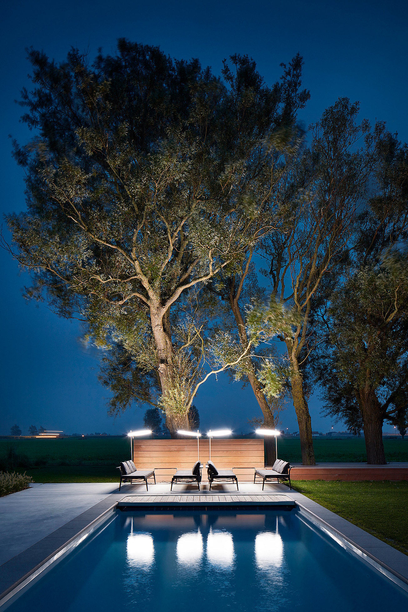 La nuit, deux transats Sol+Luna et des parasols ronds lumineux encadrent l'extrémité de la piscine, sous de grands arbres, se reflétant dans l'eau.