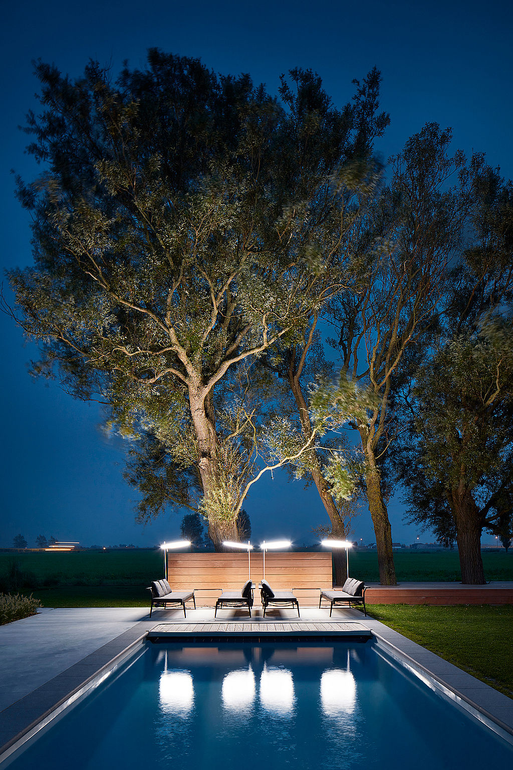 La nuit, deux transats Sol+Luna et des parasols ronds lumineux encadrent l'extrémité de la piscine, sous de grands arbres, se reflétant dans l'eau.