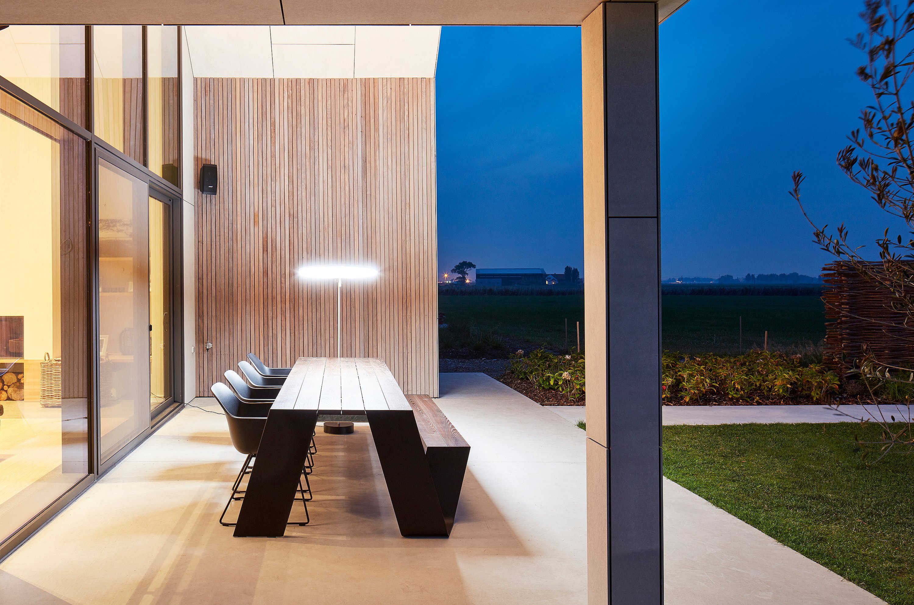 Une longue table Hopper, accompagnée de chaises longues noires, se dresse sur une terrasse couverte au crépuscule, donnant sur les champs.