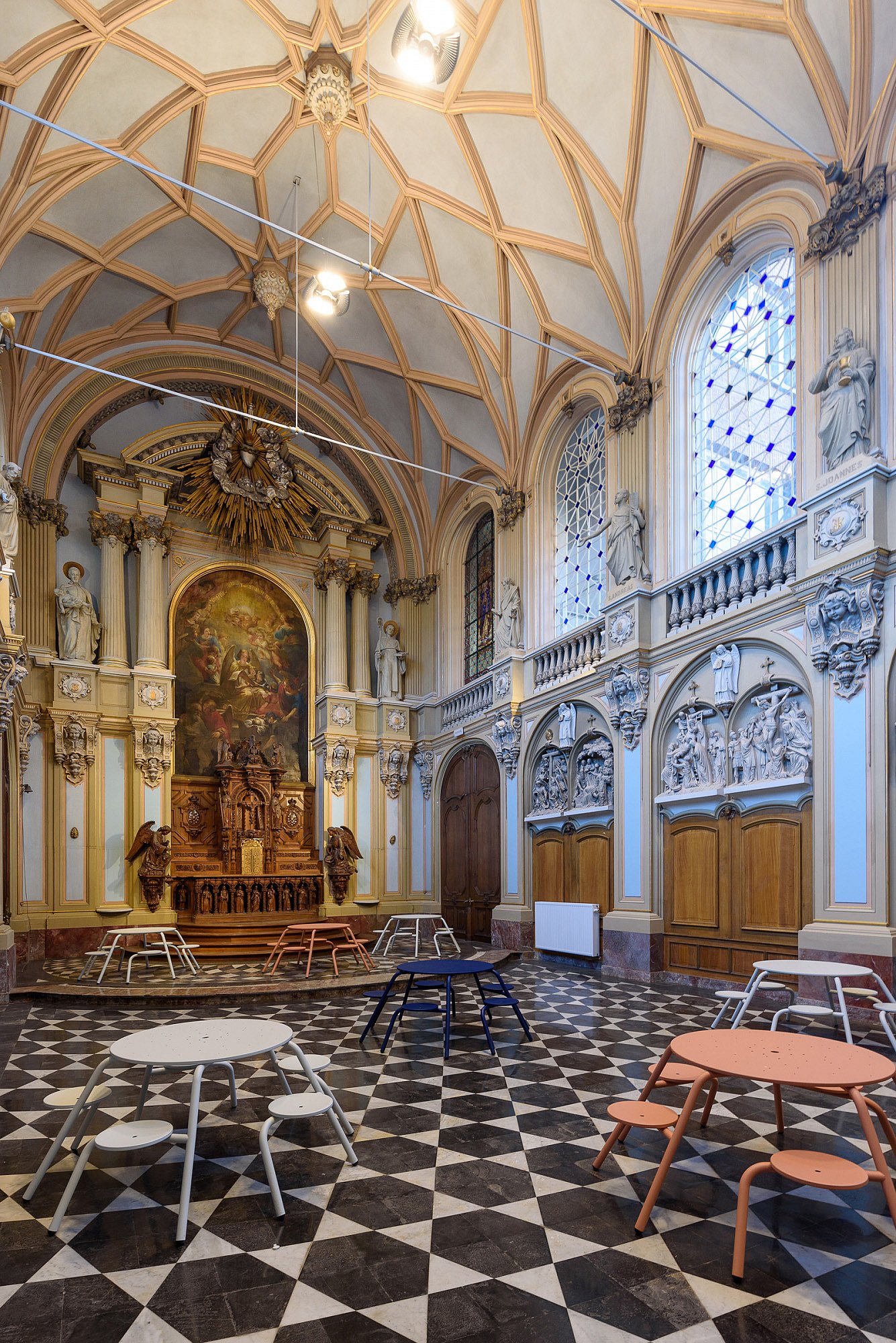 Ein restauriertes Kapellenklassenzimmer mit gewölbten Decken, Buntglasfenstern, religiösen Statuen und schwarz-weiß gefliesten Böden ist mit mehreren runden Virus-Tischen in sanften Weiß-, Blau- und Korallentönen ausgestattet.