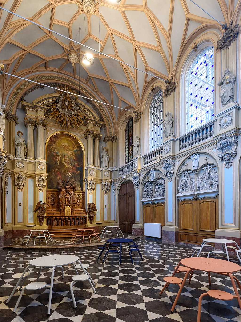 Ein restauriertes Kapellenklassenzimmer mit gewölbten Decken, Buntglasfenstern, religiösen Statuen und schwarz-weiß gefliesten Böden ist mit mehreren runden Virus-Tischen in sanften Weiß-, Blau- und Korallentönen ausgestattet.