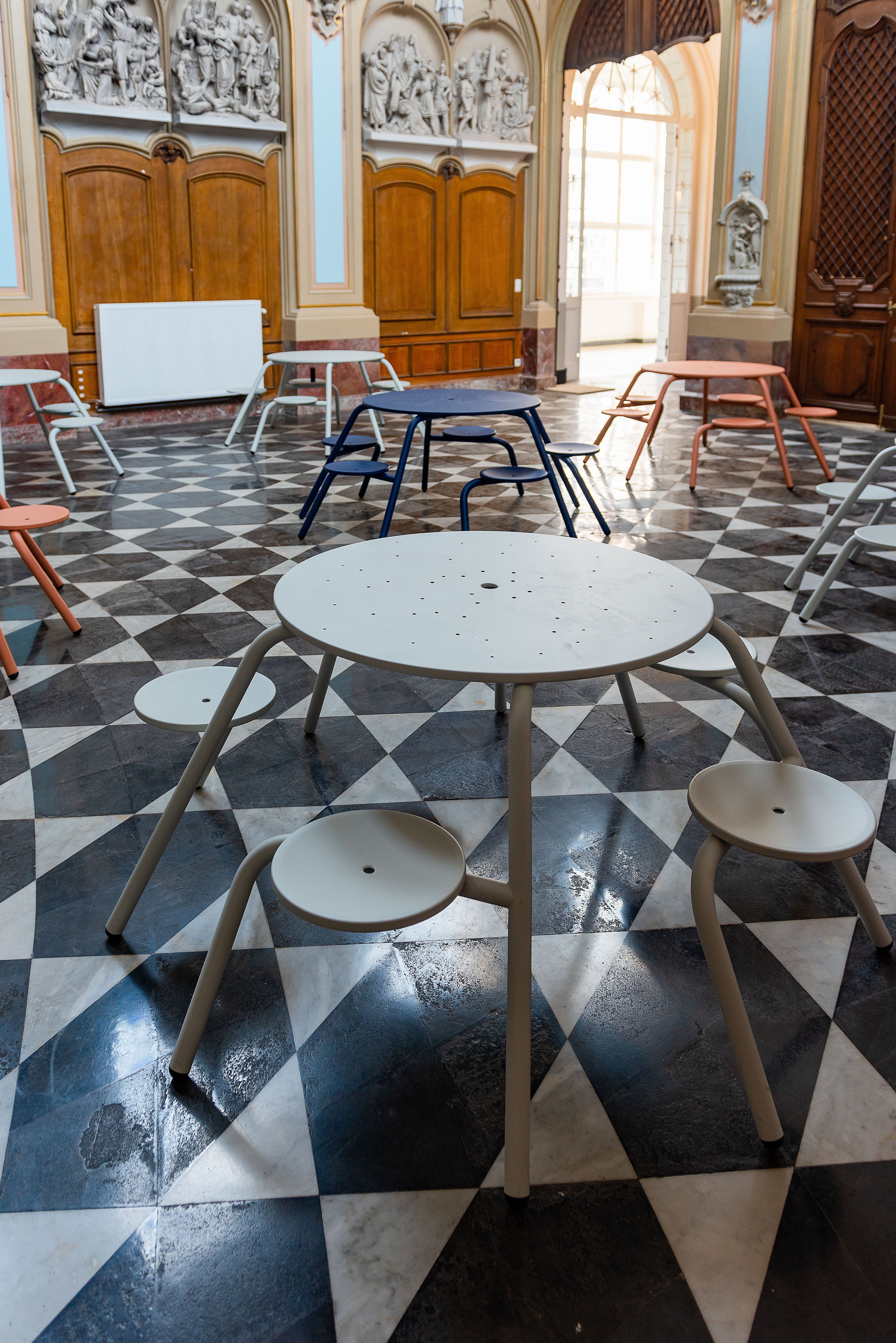 Several round Virus tables in pale grey, dark blue and coral are spread across the chapel interior, with carved wall reliefs, timber doors and sunlight entering through the open doorway.