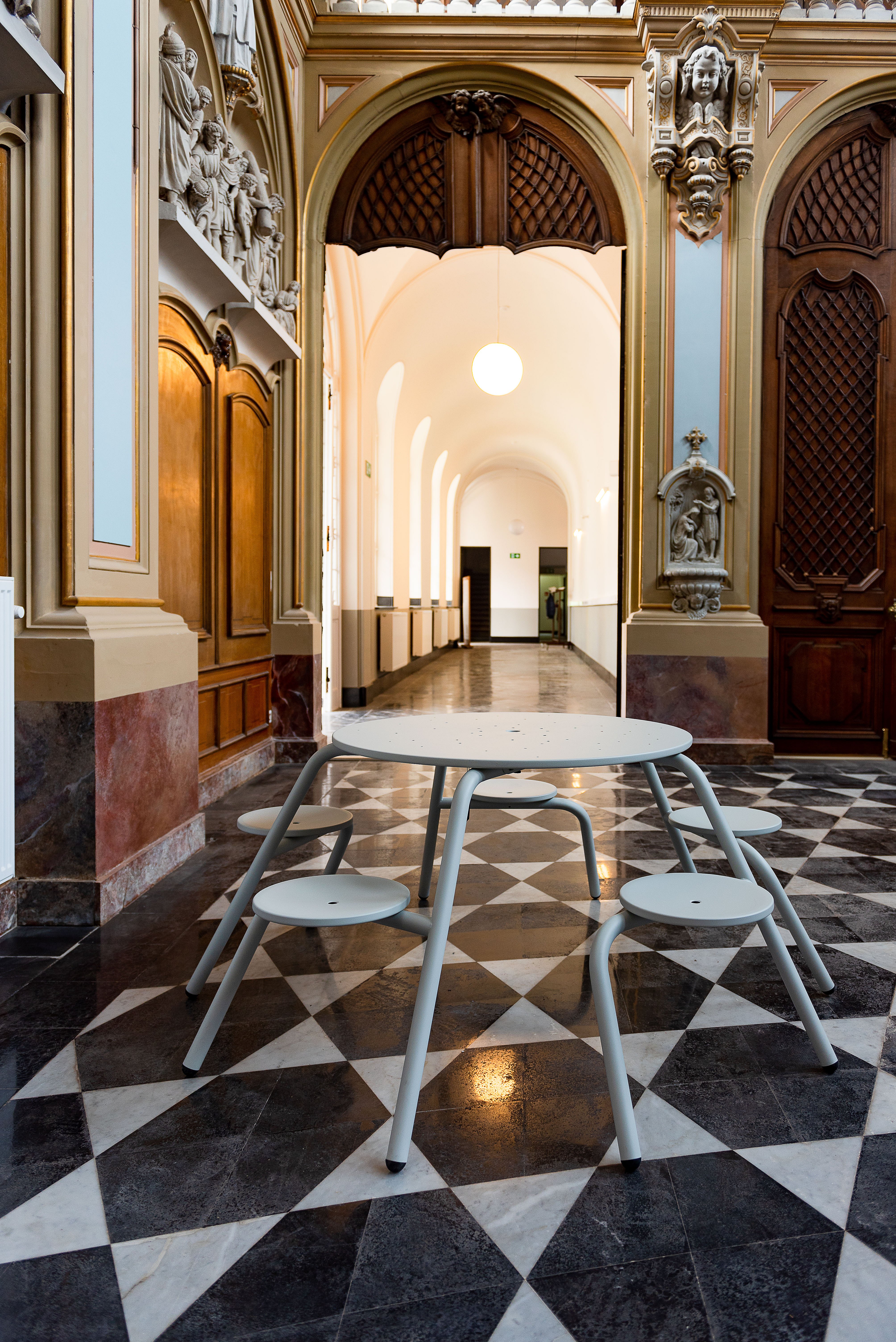A pale grey Virus table with integrated stools stands on the patterned chapel floor in front of an arched doorway, framed by carved wood panelling and ornate historic detailing.