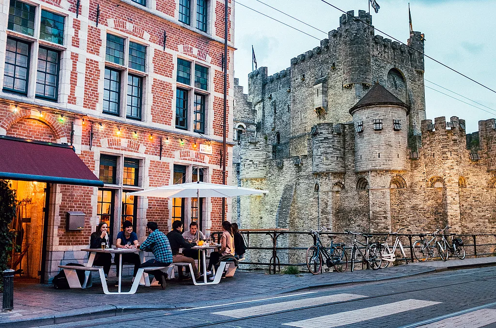 Guests dine at two white Anker picnic tables on Bodo’s canal-side terrace, with a white Inumbrina parasol and the Castle of the Counts rising beside them at dusk