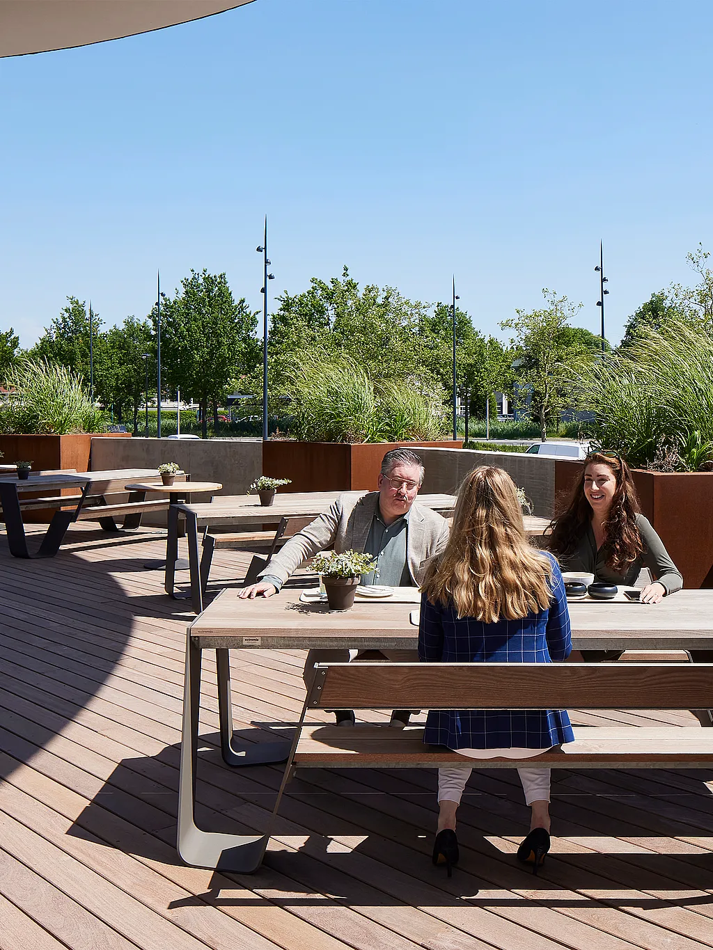 Drei Kollegen sitzen um einen Hopper-Picknicktisch aus verwittertem Holz auf der geschwungenen Dachterrasse des AFAS Experience Center, mit runden Pantagruel-Tischen und hohen Bepflanzungen dahinter