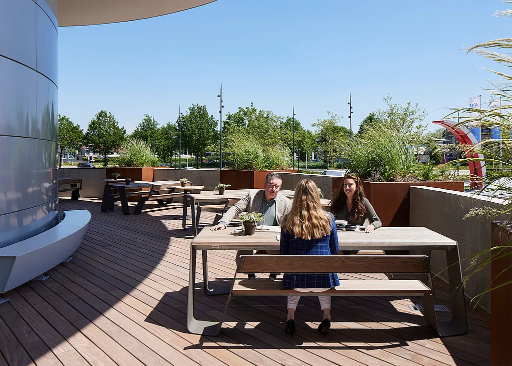 Three colleagues sit around a weathered-wood Hopper picnic table on the curved rooftop terrace of the AFAS Experience Center, with round Pantagruel tables and tall planting behind them