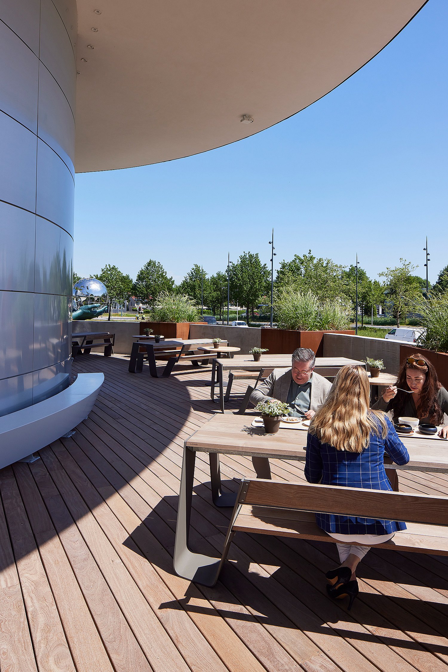 Trois collègues déjeunent à une table de pique-nique Hopper en bois vieilli sur la terrasse incurvée, avec des tables Pantagruel et des plantations réparties sur le pont derrière elles