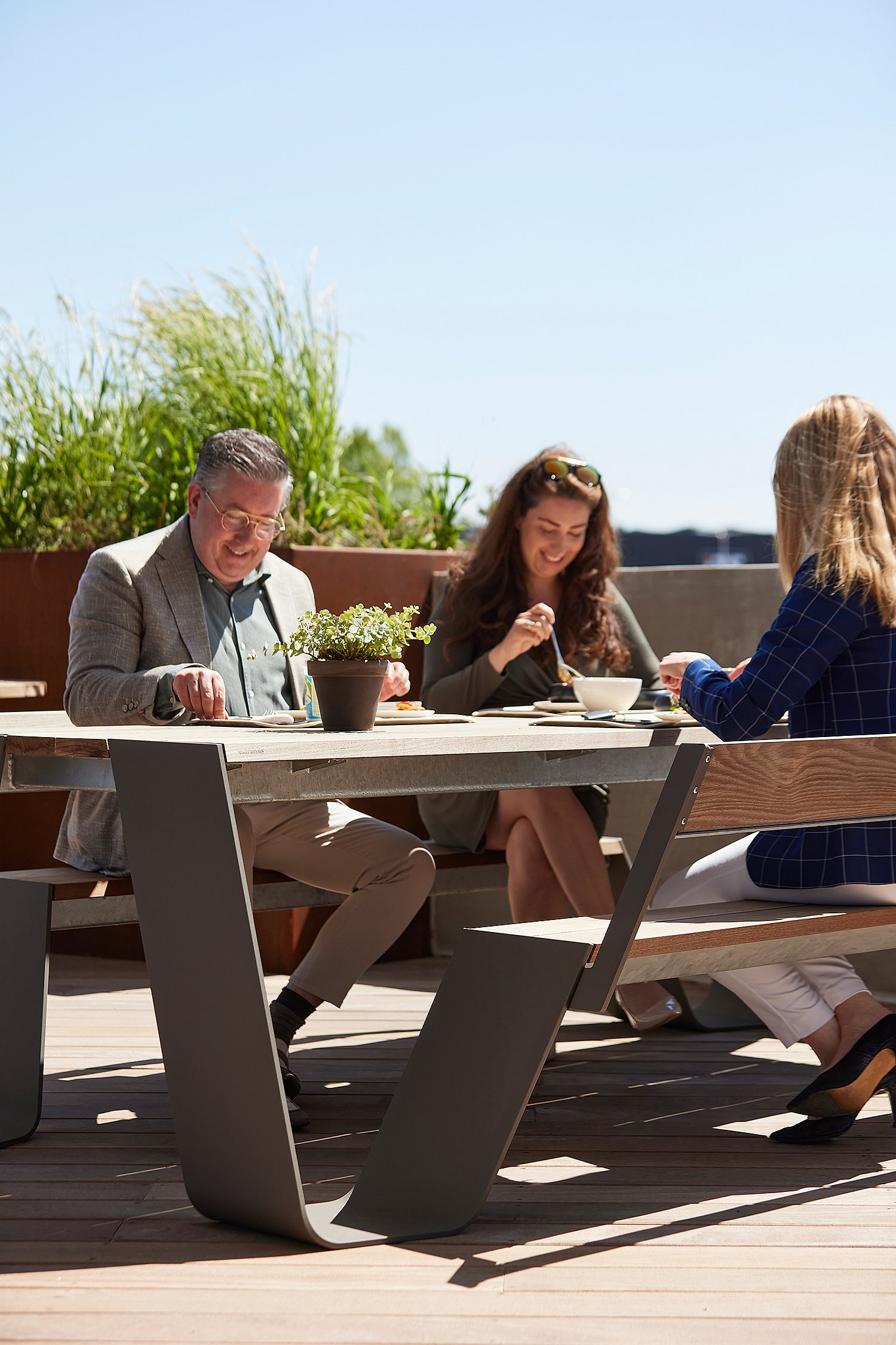 Trois collègues mangent ensemble à une table de pique-nique Hopper en bois vieilli sur la terrasse ensoleillée du toit.