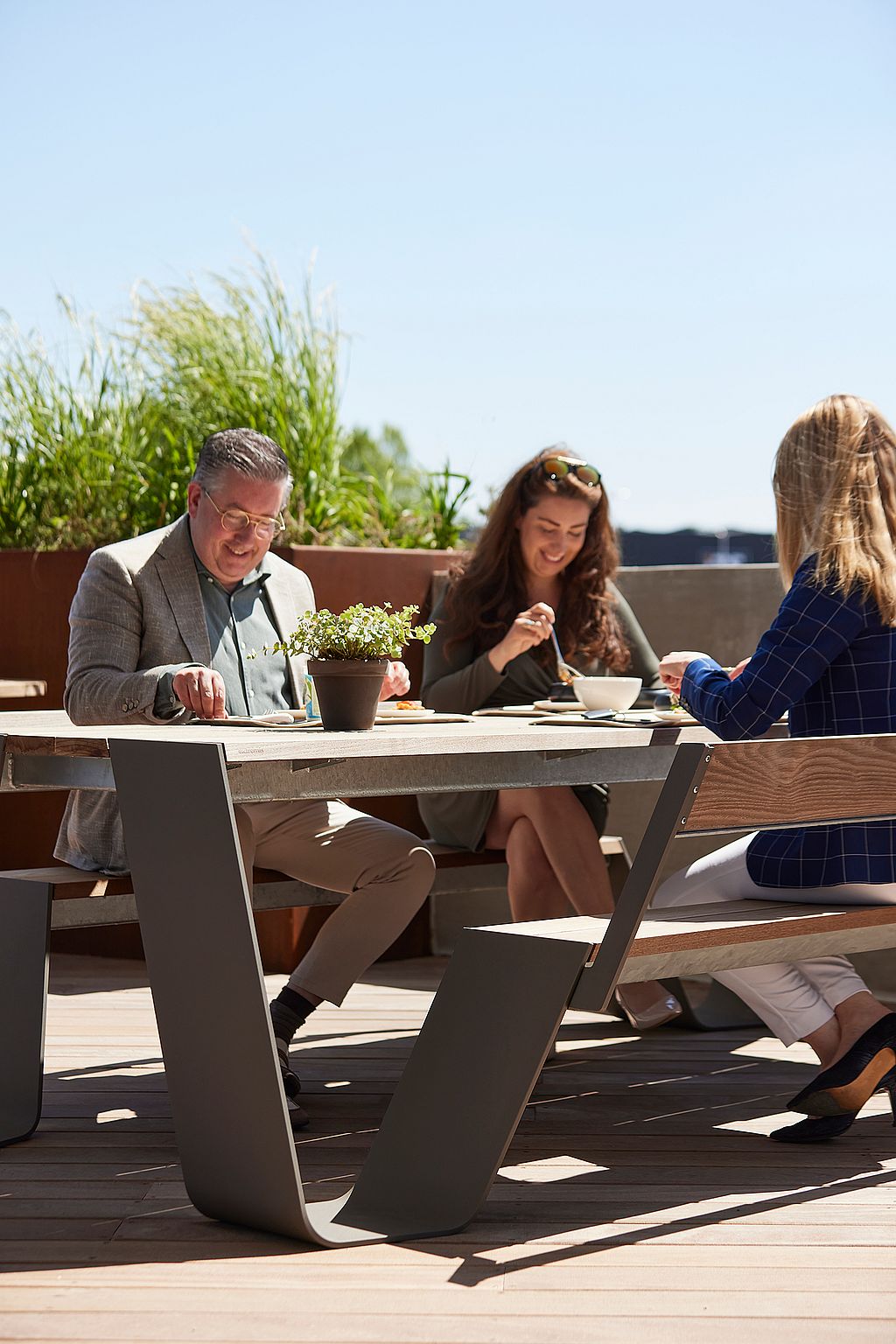 Trois collègues mangent ensemble à une table de pique-nique Hopper en bois vieilli sur la terrasse ensoleillée du toit.