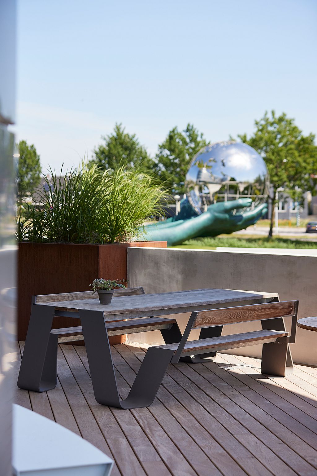 Une table de pique-nique Hopper en bois patiné se dresse sur une terrasse en bois à côté d’un mur bas en béton, avec la grande sculpture de globe en miroir visible dans le fond.
