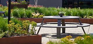 Une table de pique-nique Pantagruel grise vide se trouve parmi des jardinières rouillées et des herbes ornementales dans la cour paysagée.