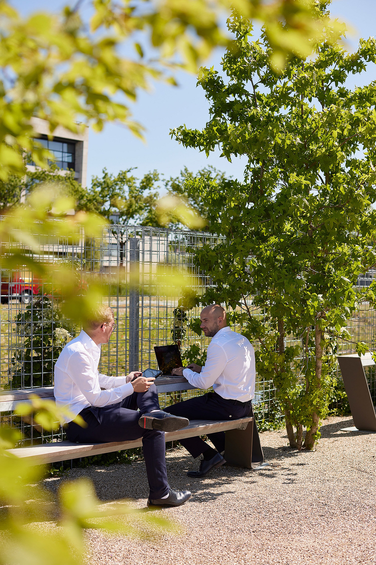 Deux collègues s’assoient sur des bancs Hopper opposés dans un espace de travail extérieur feuillu, l’un utilisant un ordinateur portable et l’autre tenant un téléphone.