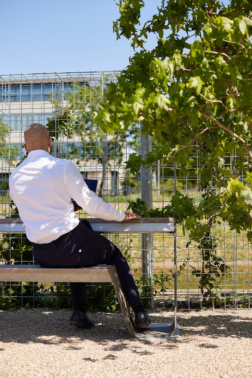 Un homme est assis sur le côté d’un banc Hopper en bois patiné, utilisant la corniche supérieure comme surface de travail sous un arbre feuillu.