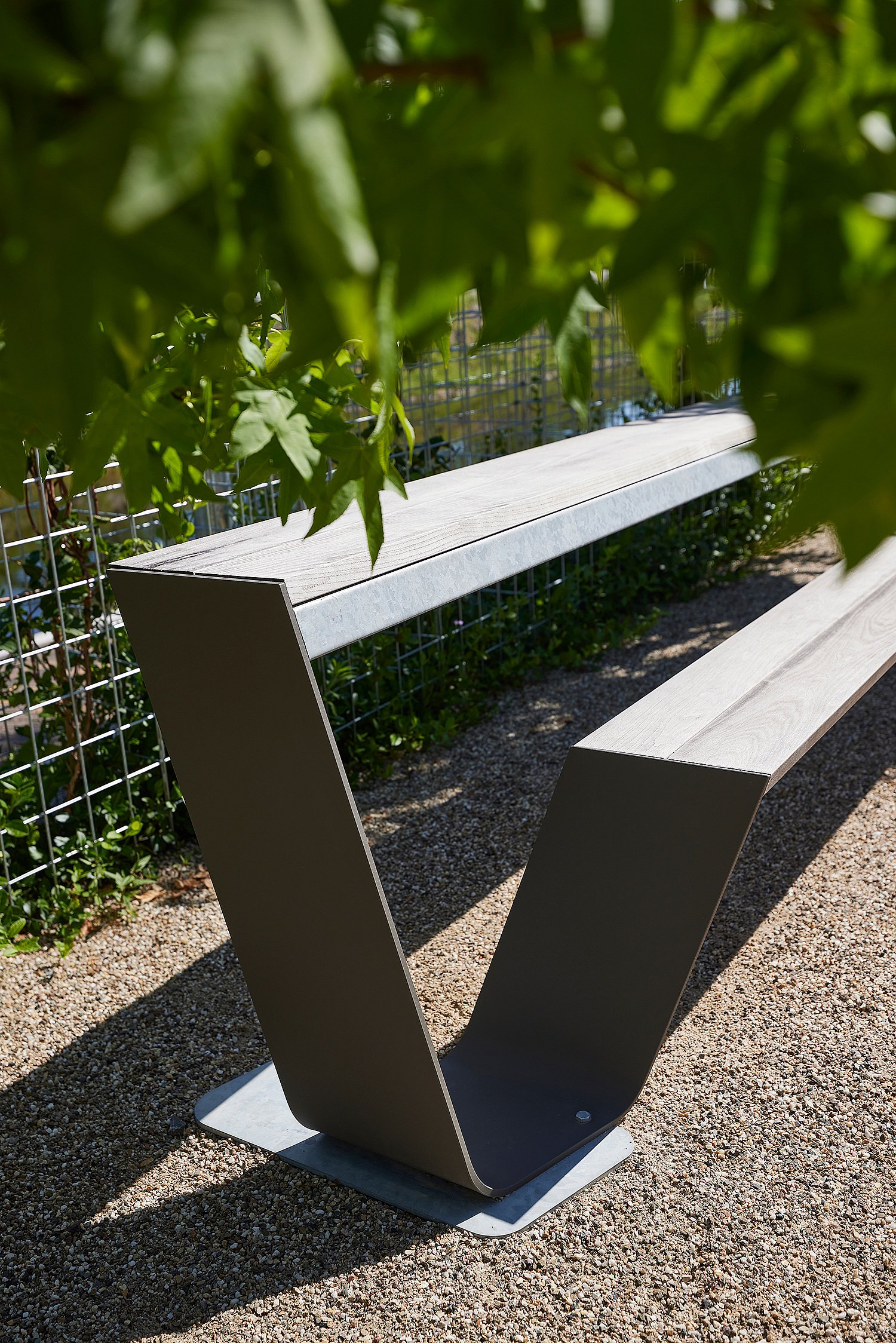 Gros plan d’un banc vide de Hopper en bois patiné sur du gravier, encadré par des feuilles vertes au premier plan.