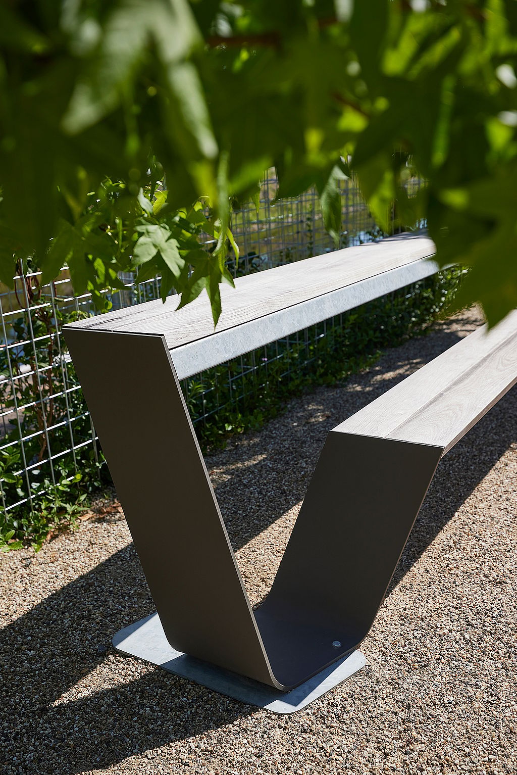 Gros plan d’un banc vide de Hopper en bois patiné sur du gravier, encadré par des feuilles vertes au premier plan.