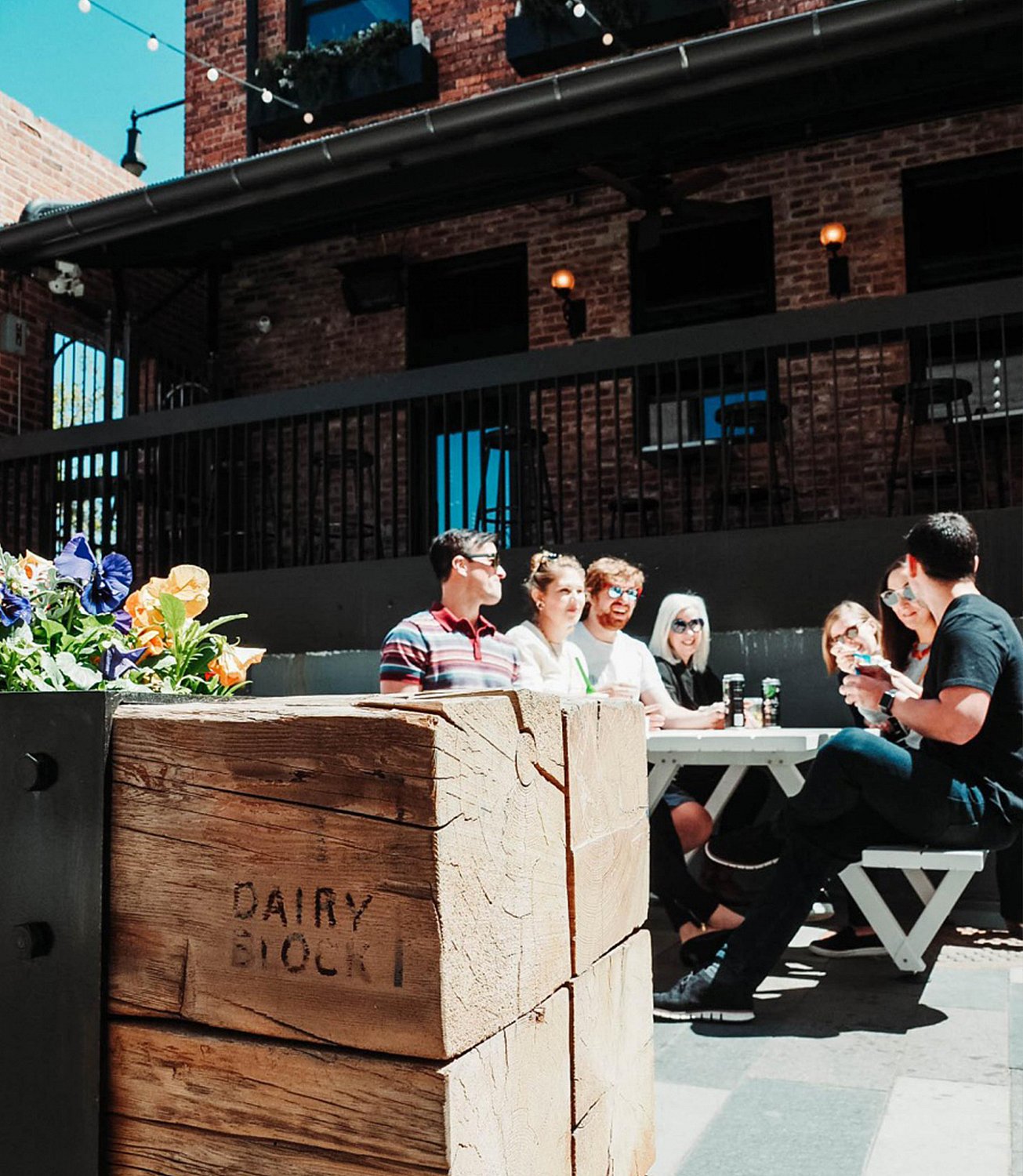 A group of friends gathers around a white Marina picnic table in the sunny Dairy Block courtyard, with a large wooden flower planter in the foreground.
