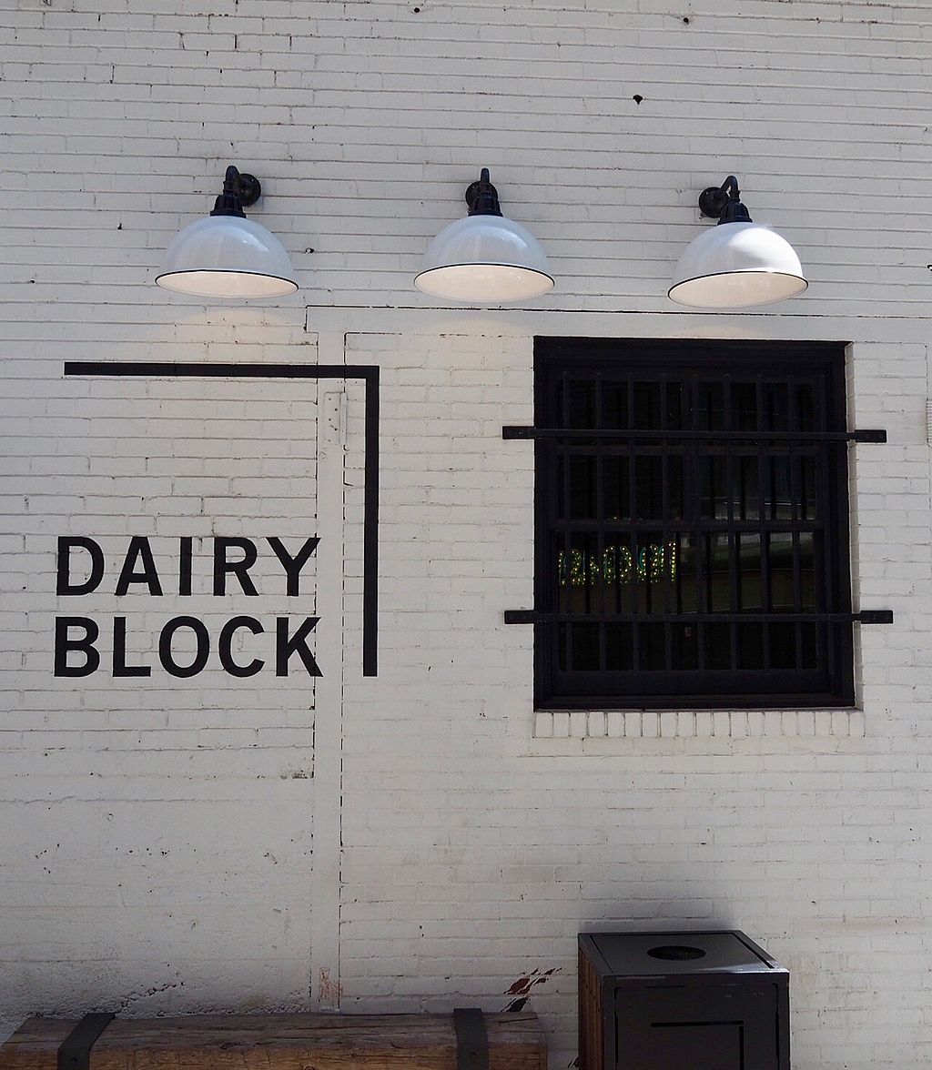 White brick wall at Dairy Block with bold black lettering, three industrial wall lamps, a barred window and a dark waste bin below.