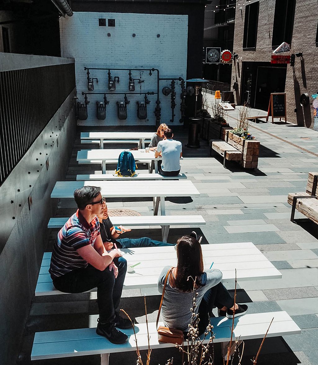 View across the courtyard showing multiple white Marina picnic tables at Dairy Block, with small groups seated in the sun and the black-pipe wall feature at the far end.