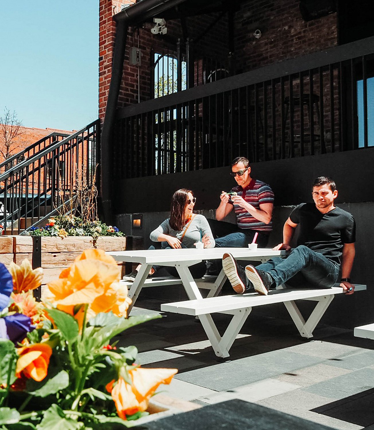 Three people relax at a white Marina picnic table beside a black railing, with bright flowers blurred in the foreground and brick walls behind.