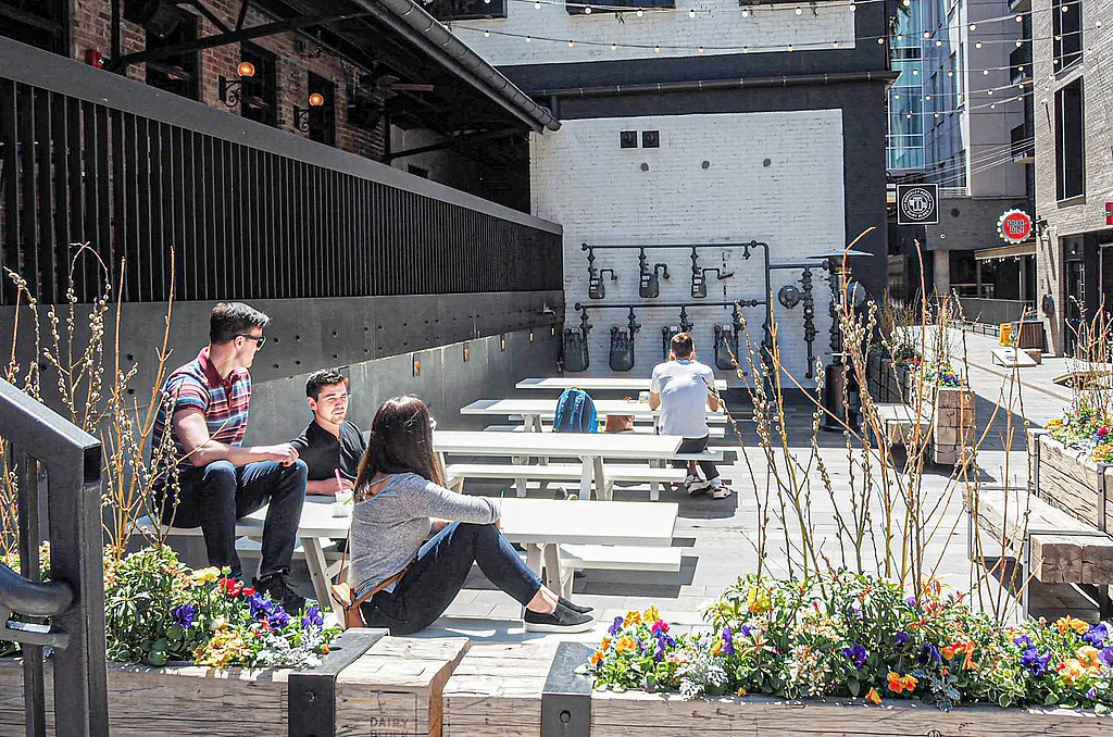 Sunken courtyard at Dairy Block with several white Marina picnic tables, where three people sit in the foreground and another person works at a table near a wall with exposed black pipes and string lights overhead.