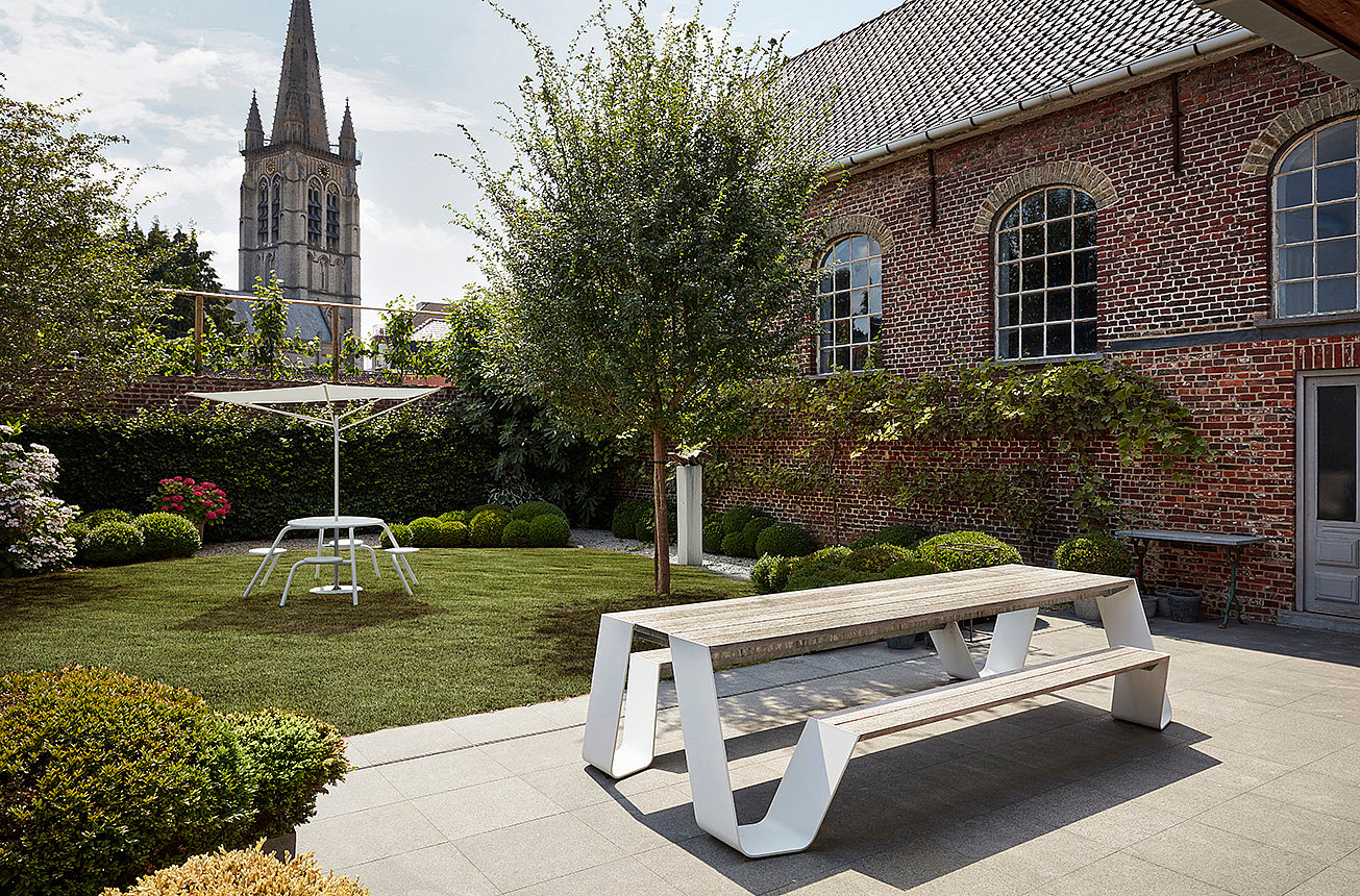 Ein Hopper-Picknick aus verwittertem Holz mit weißen Beinen steht auf einer gepflasterten Terrasse neben einer gepflegten Rasenfläche, einem Stadthaus aus Backstein und einem Kirchturm.