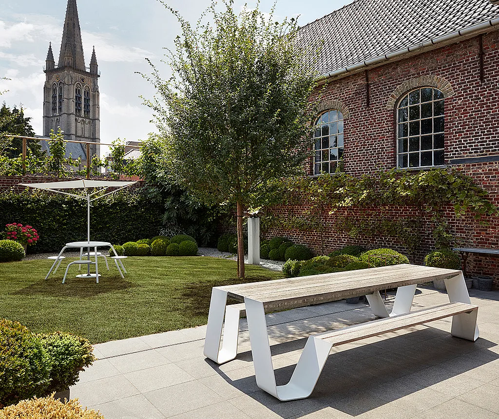 A weathered-wood Hopper picnic with white legs stands on a paved terrace beside a manicured lawn, brick townhouse and church tower.