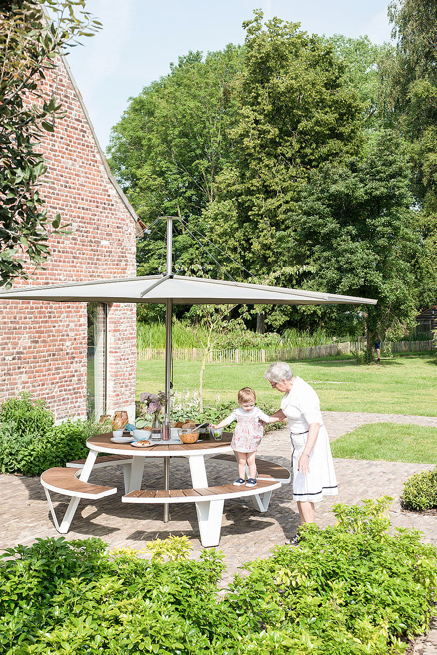 Eine ältere Frau und ein kleines Kind stehen neben einem runden Pantagruel-Tisch mit geschwungenen Holzbänken unter einem großen Inumbrina-Sonnenschirm im Gartenhof.
