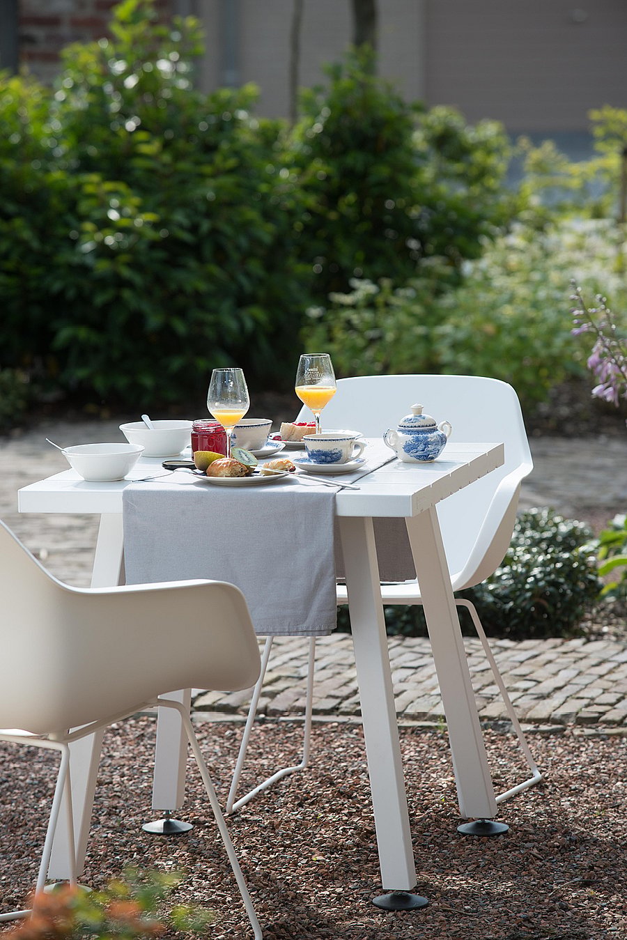 Auf dem Kieshof vor der grünen Gartenbepflanzung steht ein weißer Marina-Bistrotisch mit Orangensaft-, Tee- und Frühstücksteller.
