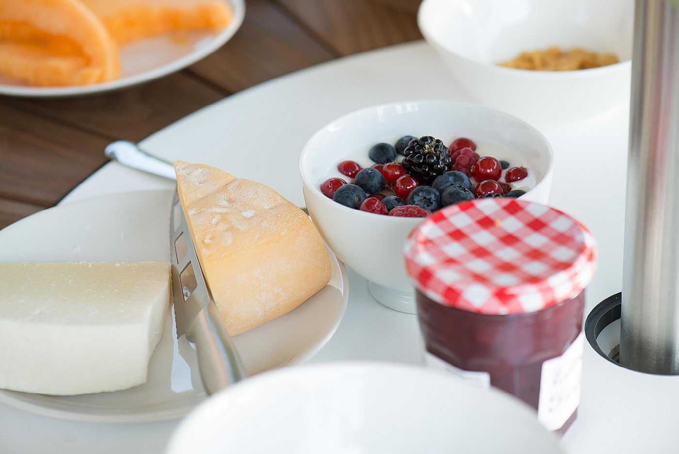 Nahaufnahme des Frühstücksessens in der weißen Mitte eines runden Pantagruel-Tisches, mit Beeren, Marmelade, Brot und Käse neben der Stange eines Inumbrina-Sonnenschirms.