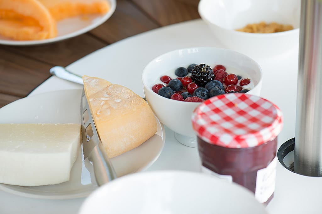 Nahaufnahme des Frühstücksessens in der weißen Mitte eines runden Pantagruel-Tisches, mit Beeren, Marmelade, Brot und Käse neben der Stange eines Inumbrina-Sonnenschirms.
