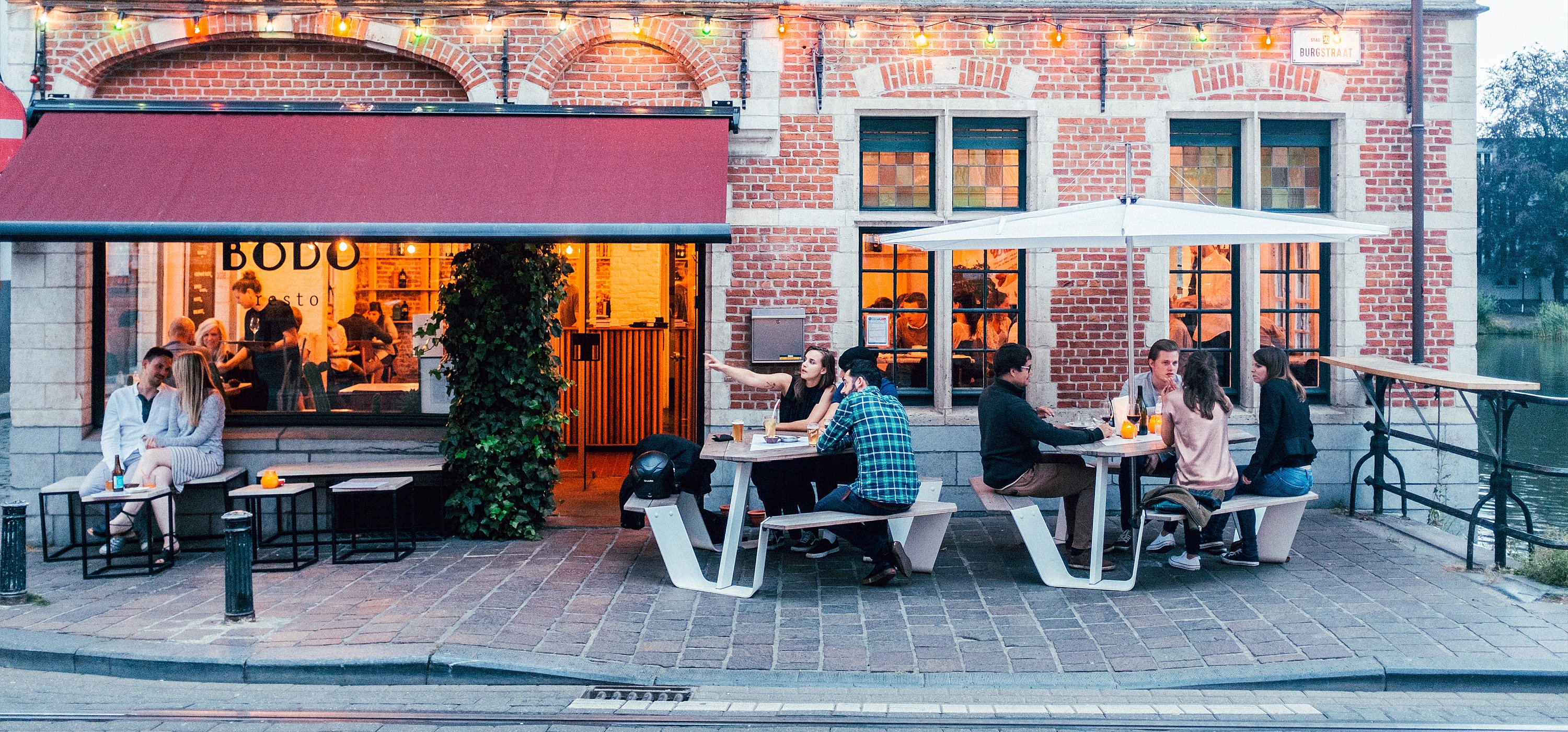 Front view of Bodo’s brick façade, where guests sit at two white Anker picnic tables under a white Inumbrina parasol on the street-side terrace.
