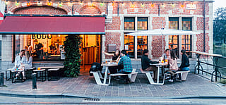 Front view of Bodo’s brick façade, where guests sit at two white Anker picnic tables under a white Inumbrina parasol on the street-side terrace.
