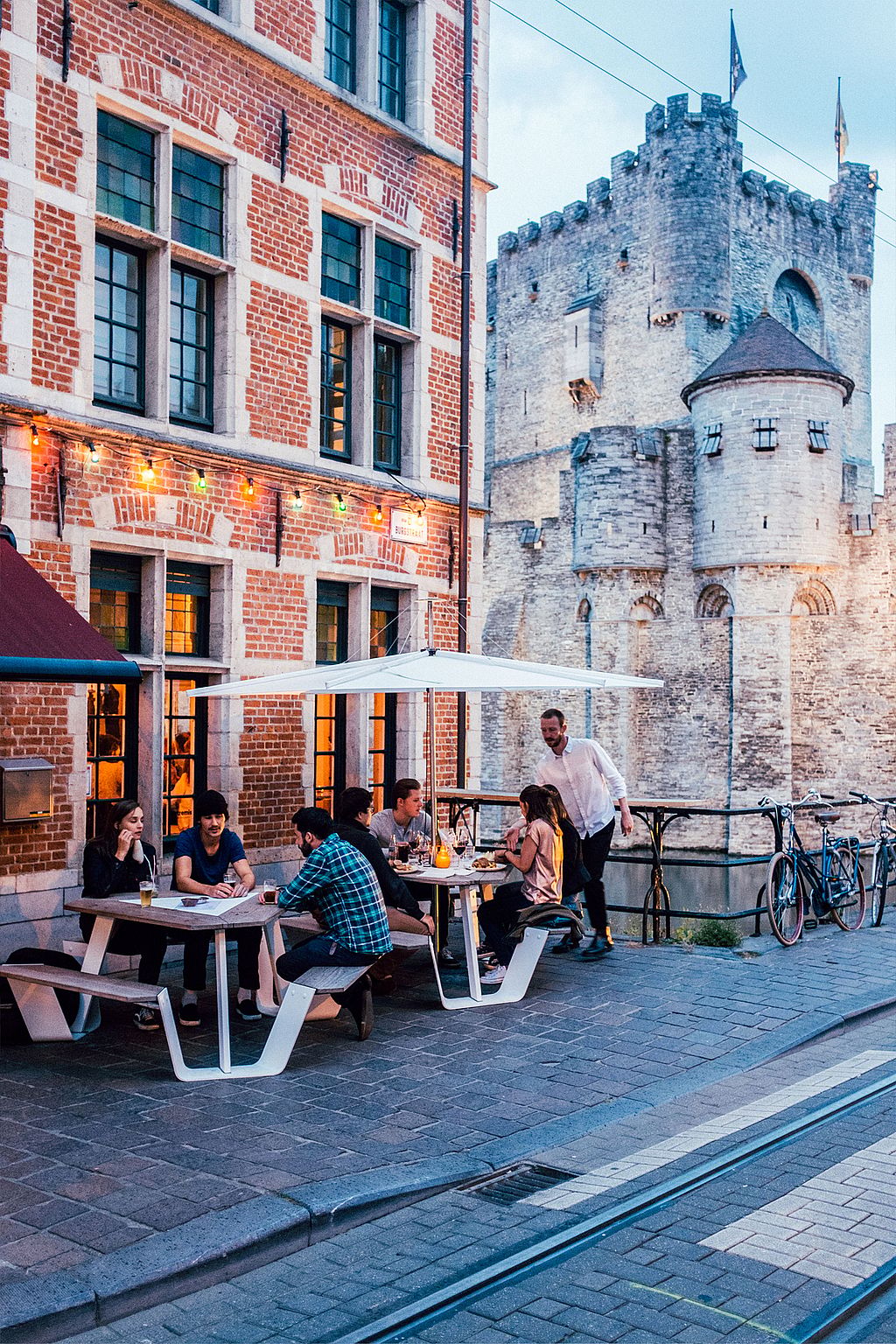 A waiter stands beside guests dining at white Anker picnic tables under a white Inumbrina parasol, with the stone walls of the castle visible behind the terrace.