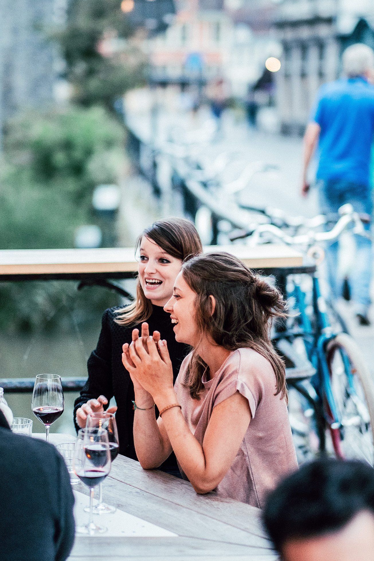 Close-up of two women chatting at an outdoor table on Bodo’s terrace, with glasses of red wine in the foreground and the canal-side railing and bicycles softly blurred behind them.
