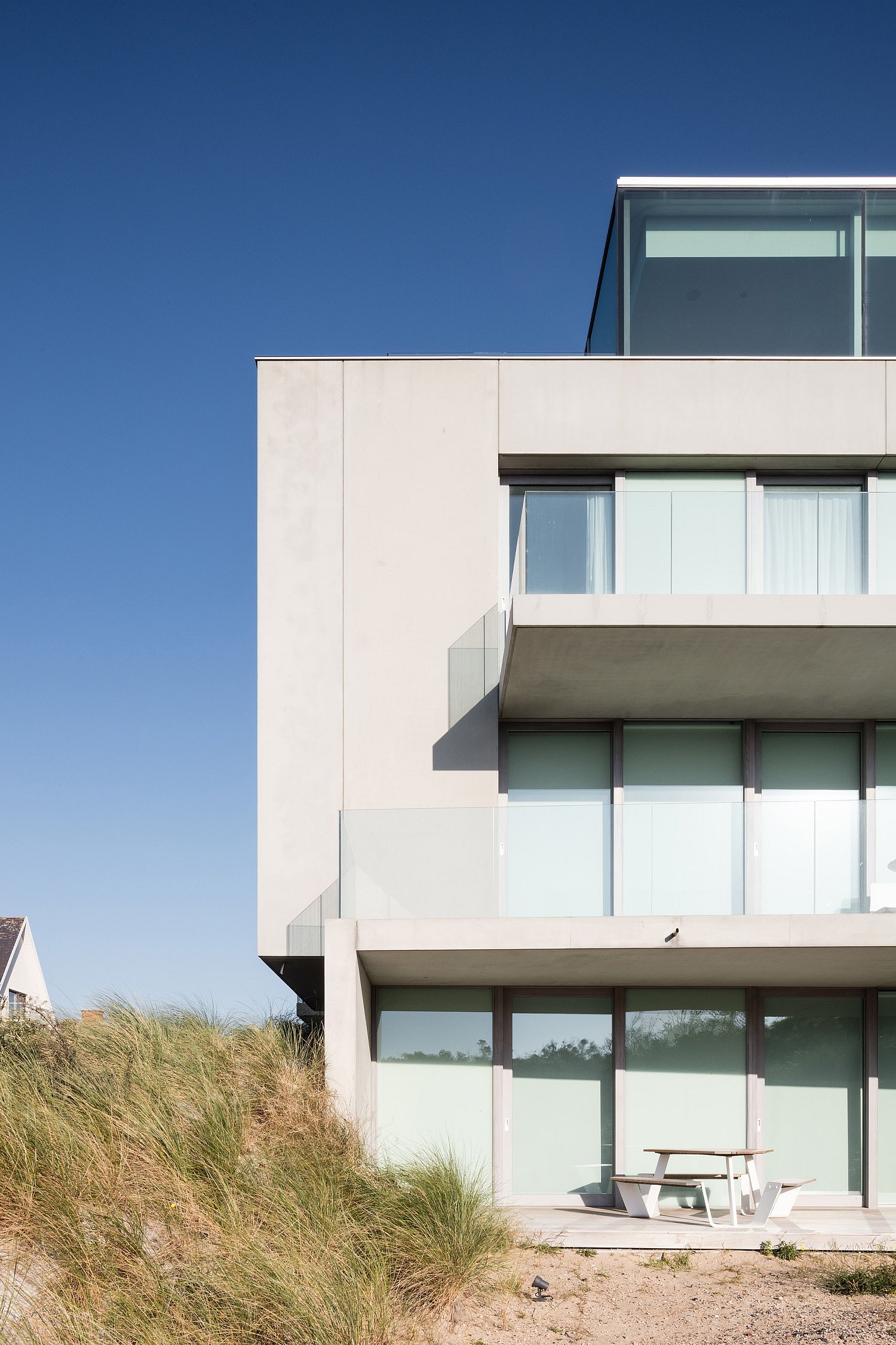 Een zijaanzicht van het moderne appartementencomplex aan zee dat boven de duinen uitsteekt, met een witte Anker picknicktafel op het terras op de begane grond onder een helderblauwe hemel.