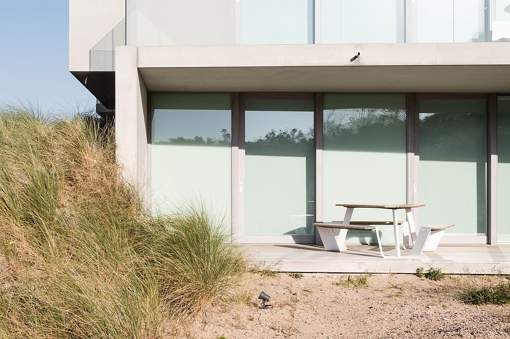 Een witte Anker picknicktafel met schuine zitjes staat op een houten terras tussen zandgrond en de glazen gevel van het appartementencomplex, omlijst door duingras.