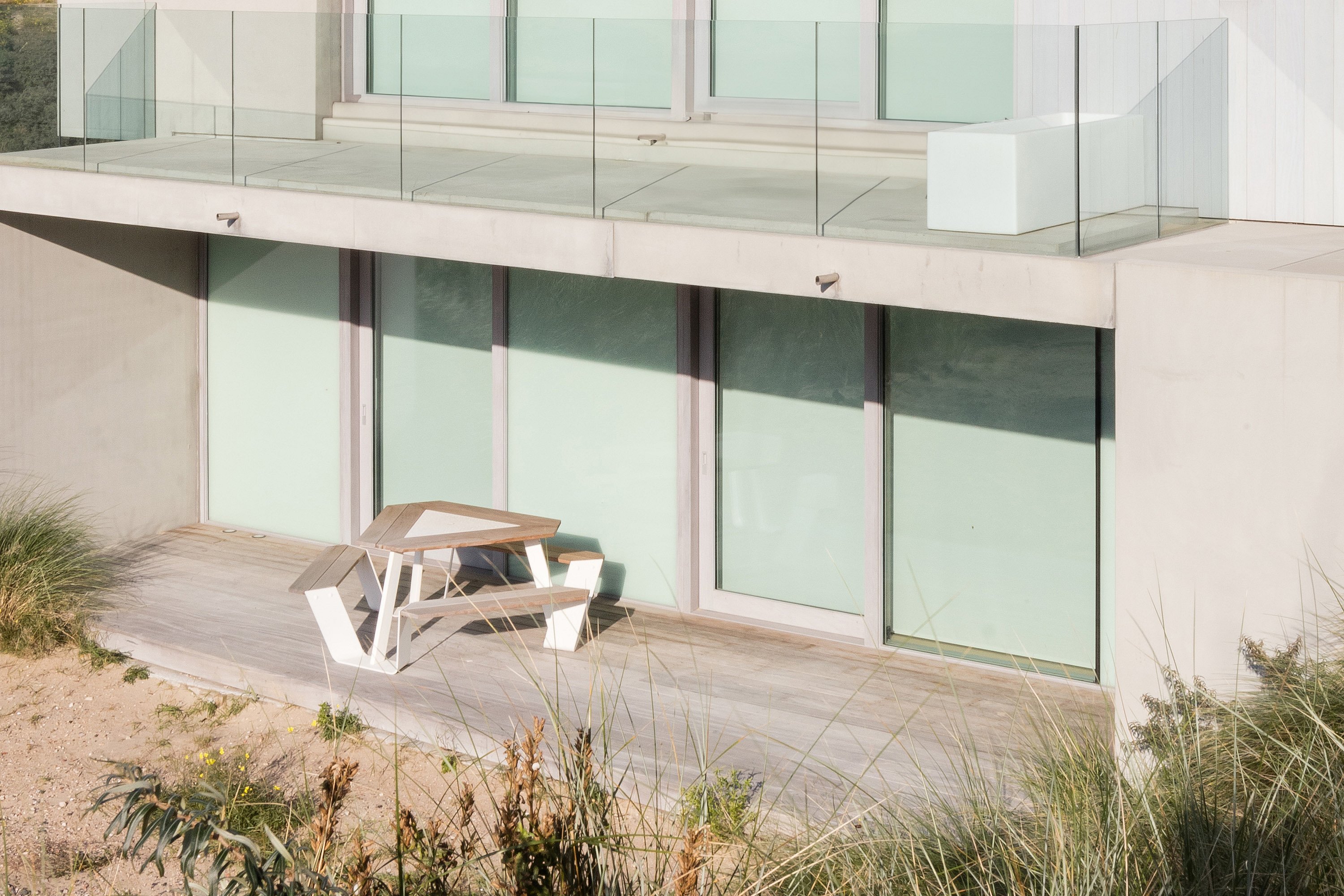 A white Anker picnic table with a wooden tabletop stands on a seaside deck in front of a modern concrete apartment building, surrounded by sandy dunes and tall beach grass.”