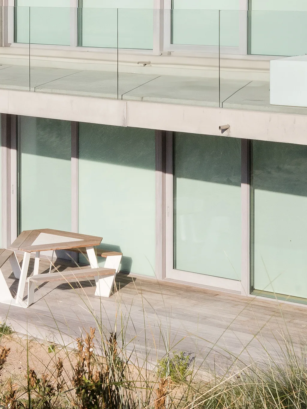 A white Anker picnic table with a wooden tabletop stands on a seaside deck in front of a modern concrete apartment building, surrounded by sandy dunes and tall beach grass.”