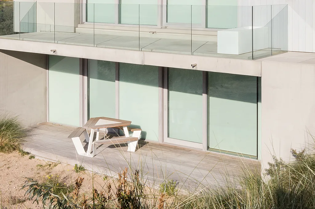 Une table de pique-nique Anker blanche, avec un plateau en bois, est installée sur une terrasse en bord de mer, devant un immeuble d'appartements moderne en béton, entourée de dunes de sable et de hautes herbes de plage.