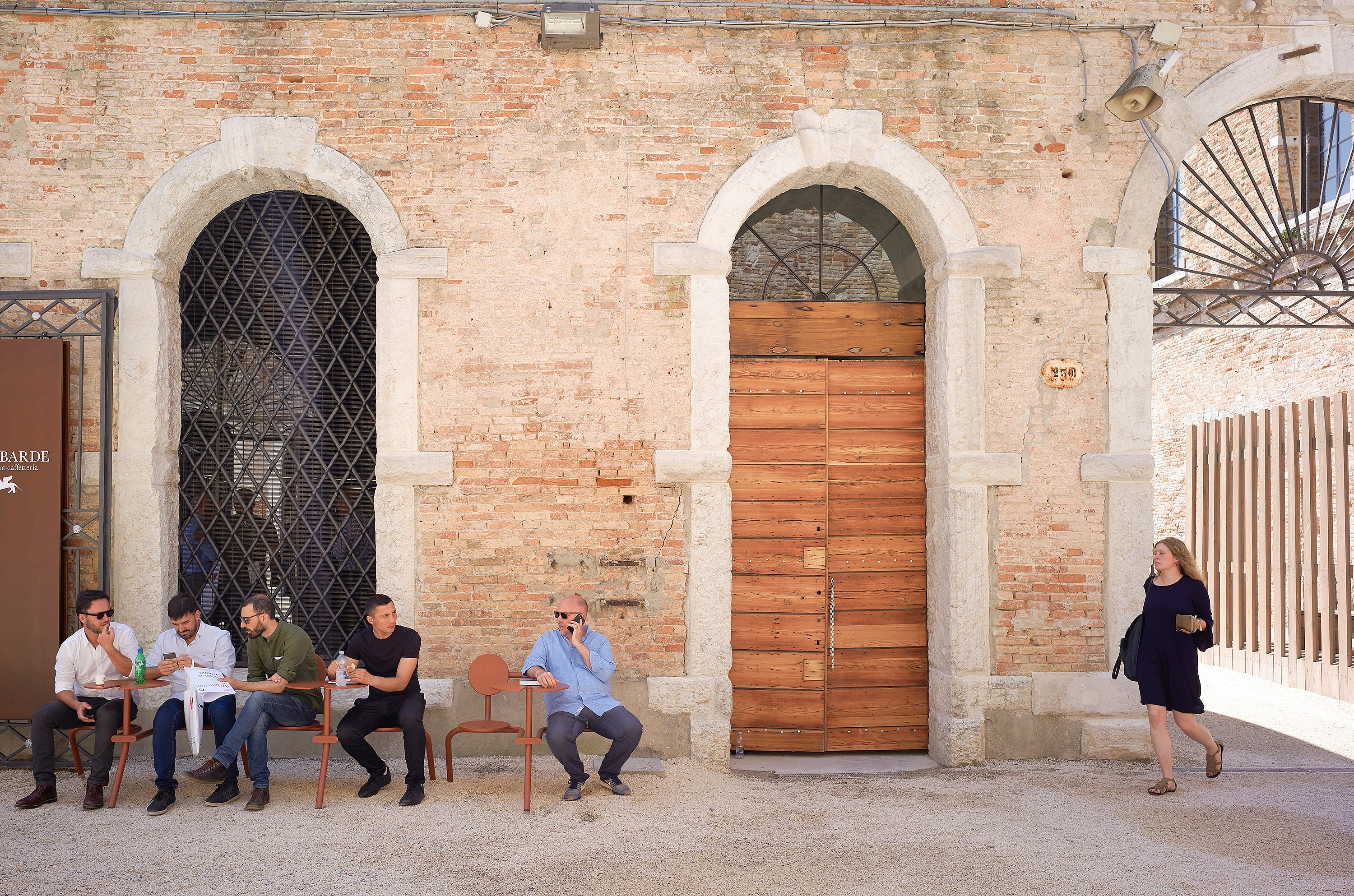 Besucher entspannen auf Bistroo-Stühlen entlang einer sonnenbeschienenen Backsteinhofmauer auf der Architekturbiennale von Venedig, mit Bogenfenstern und einer großen Holztür dahinter.