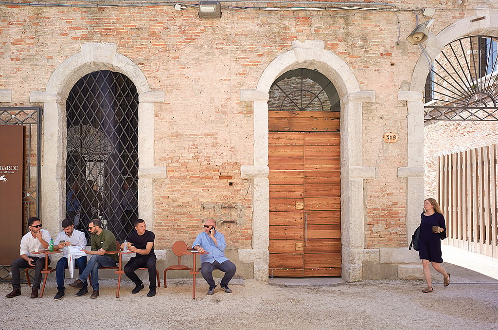 Besucher entspannen auf Bistroo-Stühlen entlang einer sonnenbeschienenen Backsteinhofmauer auf der Architekturbiennale von Venedig, mit Bogenfenstern und einer großen Holztür dahinter.