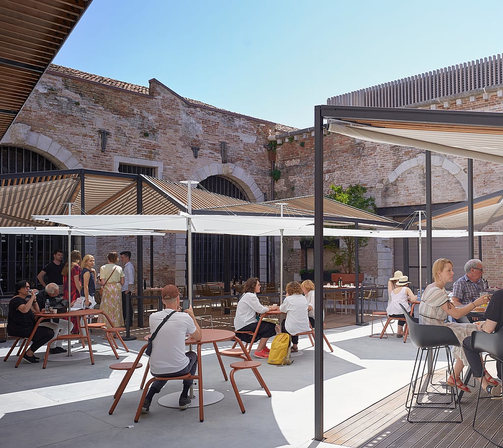 Open-Air-Hofrestaurant auf der Biennale mit Besuchern, die an Extremis-Tischen unter großen weißen Schattenüberdachungen und Regenschirmen sitzen.