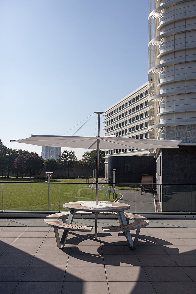 Ronde pantagrueltafel gecentreerd op het terras met een witte inumbrina paraplu boven het gazon van het ziekenhuis en de moderne gevel van het gebouw.