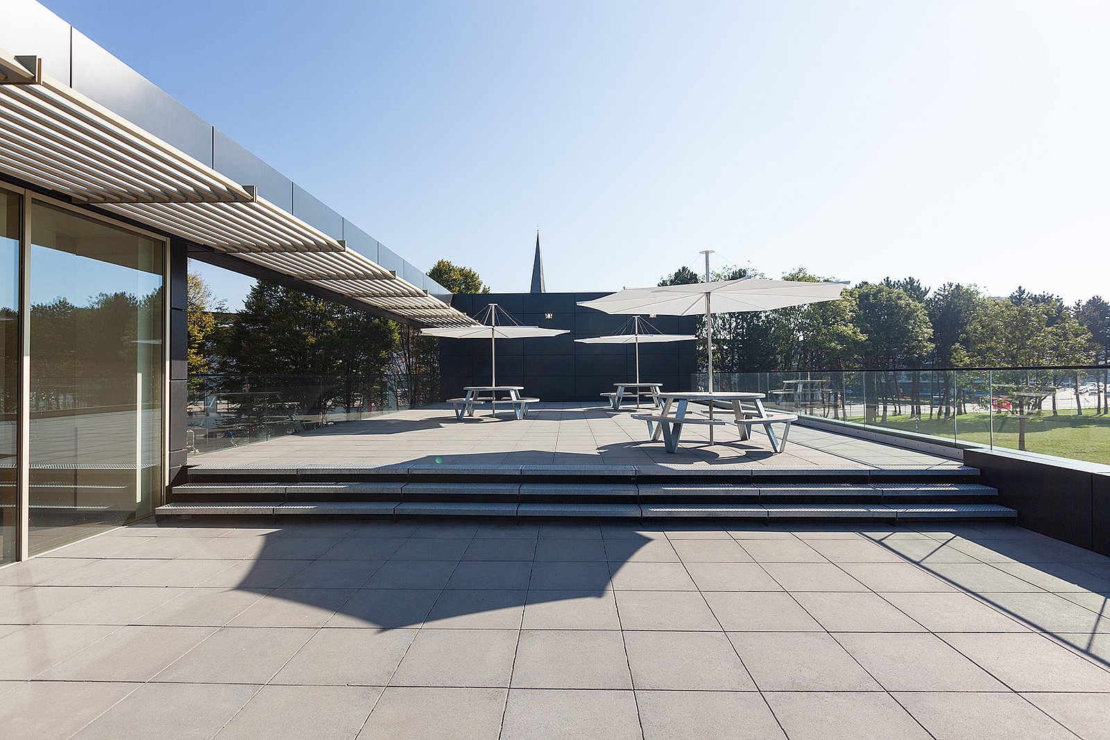 Wide view of the hospital terrace with grey paving, glass balustrades, round picnic tables and white umbrellas under a clear blue sky.