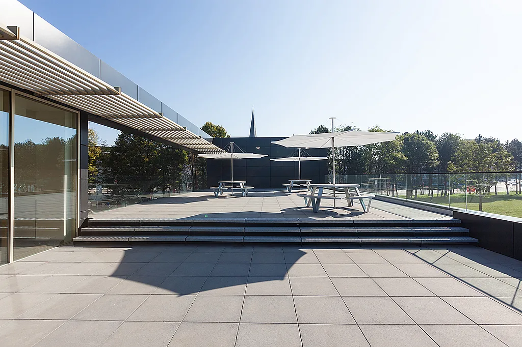 Wide view of the hospital terrace with grey paving, glass balustrades, round picnic tables and white umbrellas under a clear blue sky.