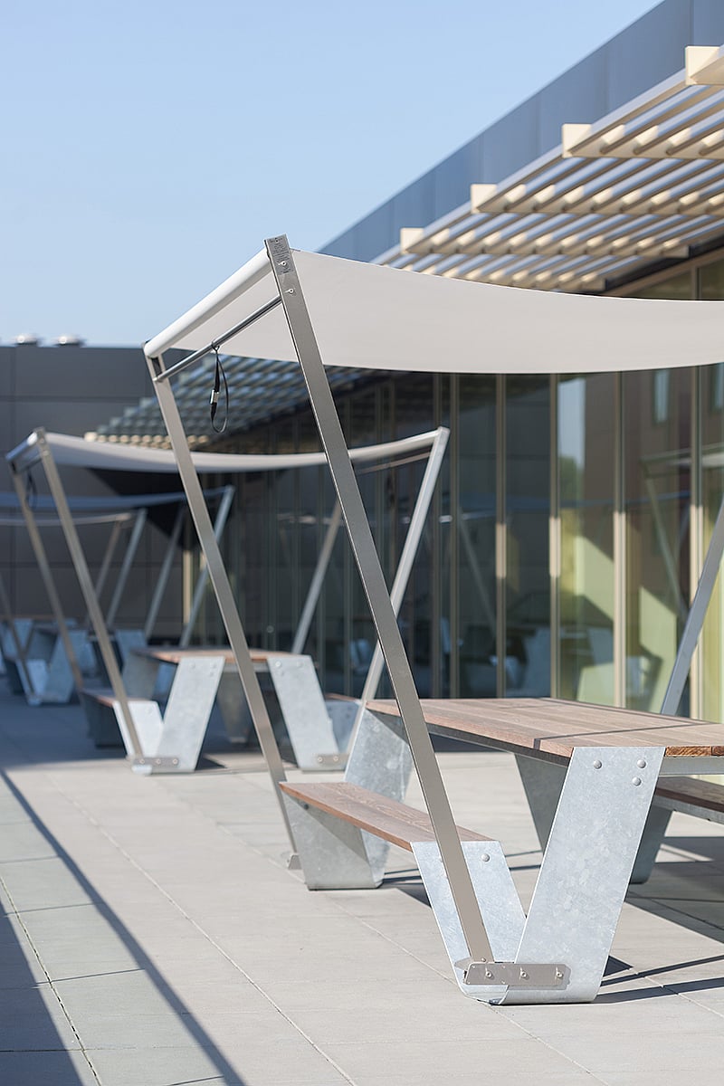 Close-up van een Hopper tafelpoot in gegalvaniseerd staal met houten banklatten, geplaatst op grote grijze terrastegels naast glazen deuren.