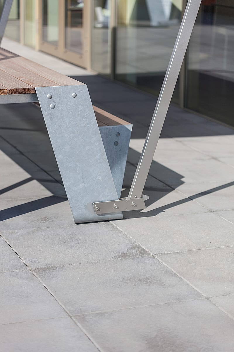Close-up van een Hopper tafelpoot in gegalvaniseerd staal met houten banklatten, geplaatst op grote grijze terrastegels naast glazen deuren.