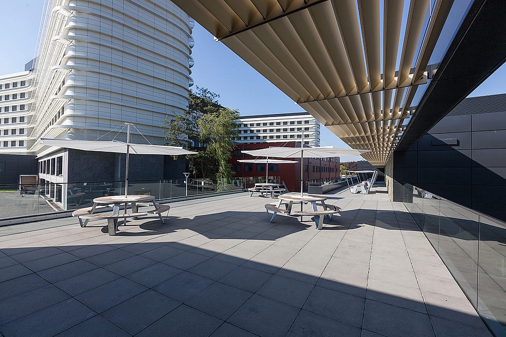 Lang dakterras naast het ziekenhuisgebouw met ronde picknicktafels, witte parasols, glazen leuningen en gebogen witte gevels op de achtergrond.