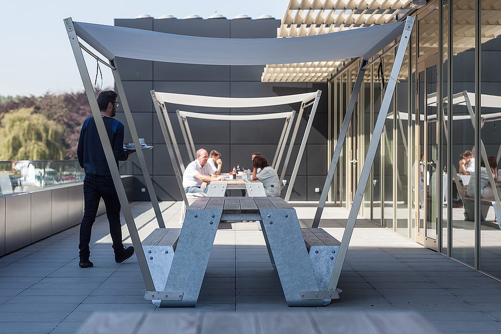 Perspectief uitzicht door een schaduwrijke hopper tafelluifel op het terras van het ziekenhuis, met personeel op de achtergrond en een persoon die voorbij loopt.